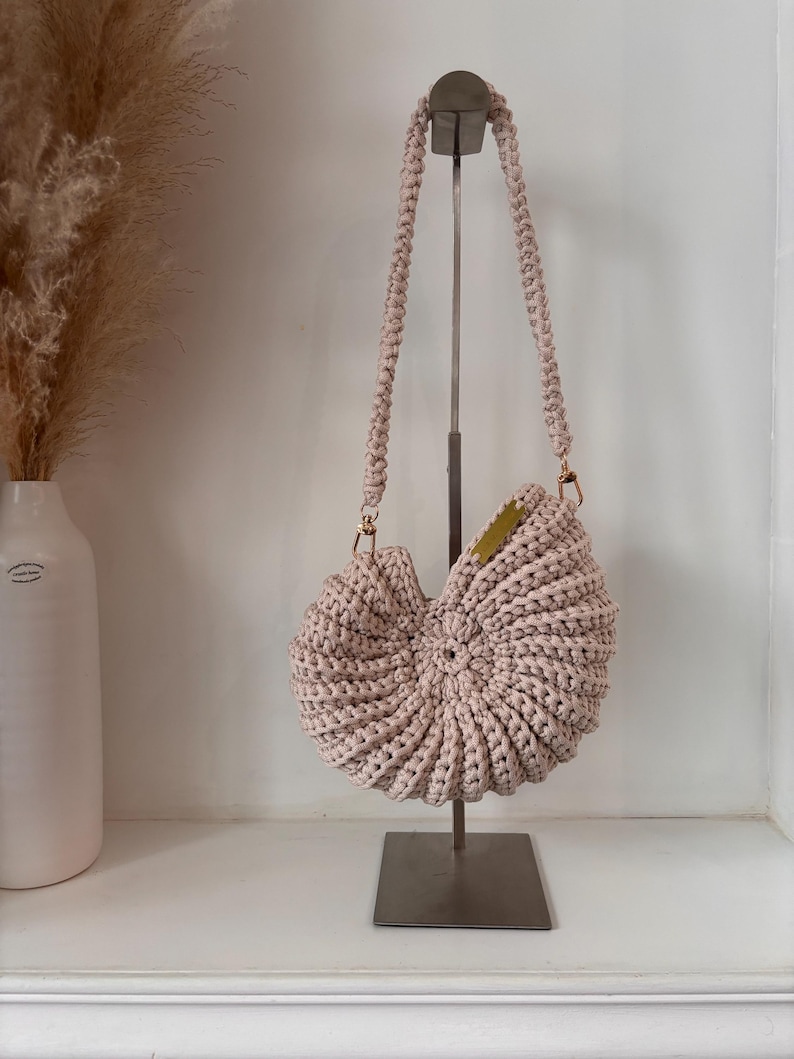 Op de afbeelding: Een lichtbeige gehaakte handtas met een opvallende spiraalvormige schelpvorm. De tas heeft een dikke, gevlochten schouderband bevestigd met goudkleurige metalen hardware en een klein rechthoekig goudkleurig metalen label aan de rechterbovenrand.