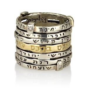 Puede incluir: Un anillo de plata y oro con múltiples bandas, cada una inscrita con texto hebreo. El anillo está engastado con dos pequeños diamantes.