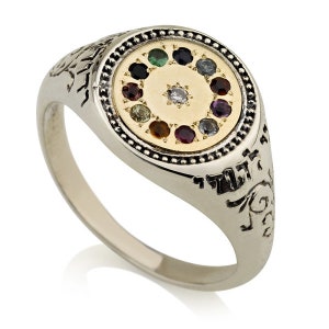 Pode incluir: Um anel de prata com um centro de ouro que apresenta um círculo de doze pedras preciosas coloridas que rodeiam um único diamante branco. O anel é gravado com texto hebraico.