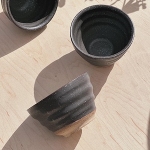May include: Three black ceramic bowls with a speckled interior and a textured, ridged exterior. The bowls are arranged on a light-colored wooden surface, with shadows from foliage adding depth. The bowls have a rustic, handcrafted appearance.