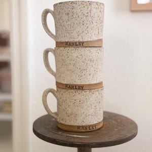 Op de afbeelding: Drie gestapelde, gespikkelde keramische mokken met gebogen handvatten. Elke mok heeft een bruine band met de naam "MANLEY" erop gedrukt. De mokken zijn beige met bruine spikkels en staan op een donker metalen oppervlak.