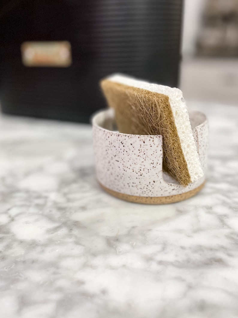 Op de afbeelding: Een witte keramische sponshouder met bruine vlekken en een bruine en witte spons erin.