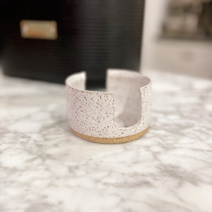 Op de afbeelding: Een witte keramische zeepbak met een gespikkelde afwerking en een gouden rand. De zeepbak heeft een uitsparing in het midden voor een stuk zeep.