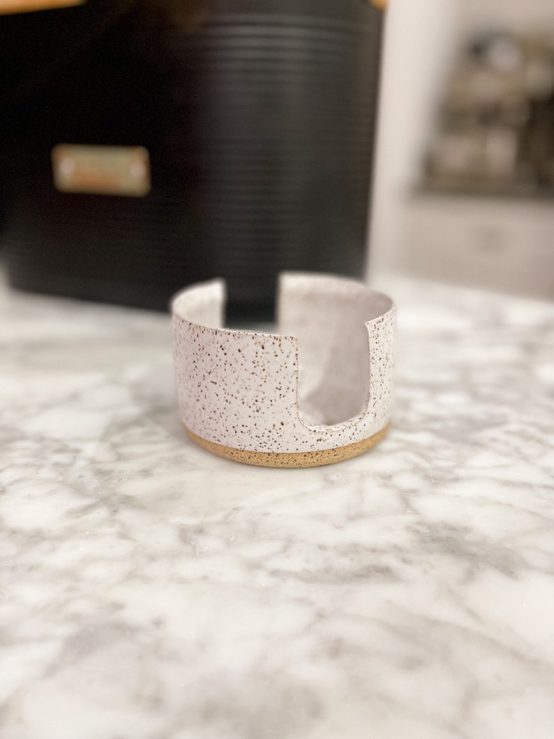 Op de afbeelding: Een witte keramische zeepbak met een gespikkelde afwerking en een gouden rand. De zeepbak heeft een uitsparing in het midden voor een stuk zeep.