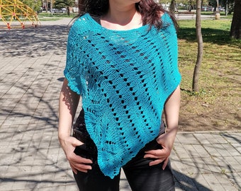 Puede incluir: Un poncho de crochet turquesa con un patrón de encaje. El poncho se lleva sobre una camisa negra y unos vaqueros.