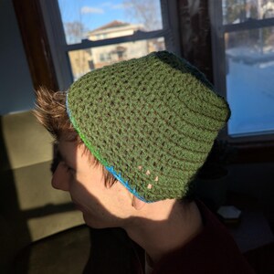 Puede incluir: Un gorro de punto de color verde con ribete azul y verde. El gorro está puesto en la cabeza de una persona.