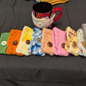 May include: A collection of colorful, crocheted mug cozies in various hues, including green, orange, yellow, blue, pink, and gray. Each cozy features a button closure and is designed to fit around a mug. A red mug with a floral cozy is also present.