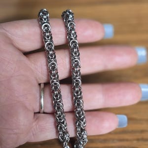 May include: A silver-colored Byzantine chain bracelet. The bracelet is made of interlocking metal rings, creating a textured, woven appearance. The clasp is visible at the end of the bracelet. The bracelet is displayed against a wooden background.