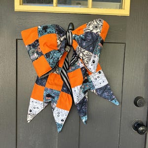 May include: A large, decorative bow made of patchwork fabric, featuring orange, teal, and patterned squares with Halloween motifs. The bow is hanging on a dark gray door, creating a festive seasonal decoration.