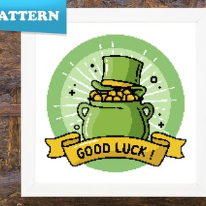 Puede incluir: Una olla de oro verde con un sombrero de duende en la parte superior. La olla está rodeada de un círculo verde con rayos blancos y las palabras "¡Buena suerte!" en una pancarta amarilla.