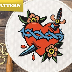 Könnte beinhalten: Ein Kreuzstichmuster mit einem roten Herzen mit einem blauen Band, das von einem Dolch durchbohrt wird und von Dornen und Blumen umgeben ist. Das Herz blutet rote Tropfen.