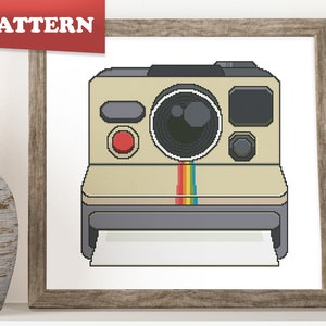 May include: A cross-stitch pattern of a vintage Polaroid camera with a rainbow stripe across the front. The camera is beige with a black lens and a black film compartment.