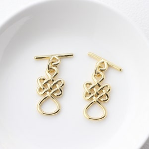 Può includere: Due chiusure a scatto dorate con un design a nodo. Le chiusure sono su uno sfondo bianco.