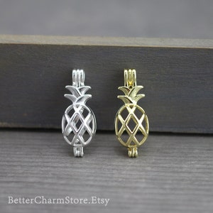 Könnte beinhalten: Zwei ananasförmige Anhängerkäfige, einer silber und einer gold. Die Käfige sind offen und bereit, eine Perle oder einen anderen kleinen Anhänger aufzunehmen.