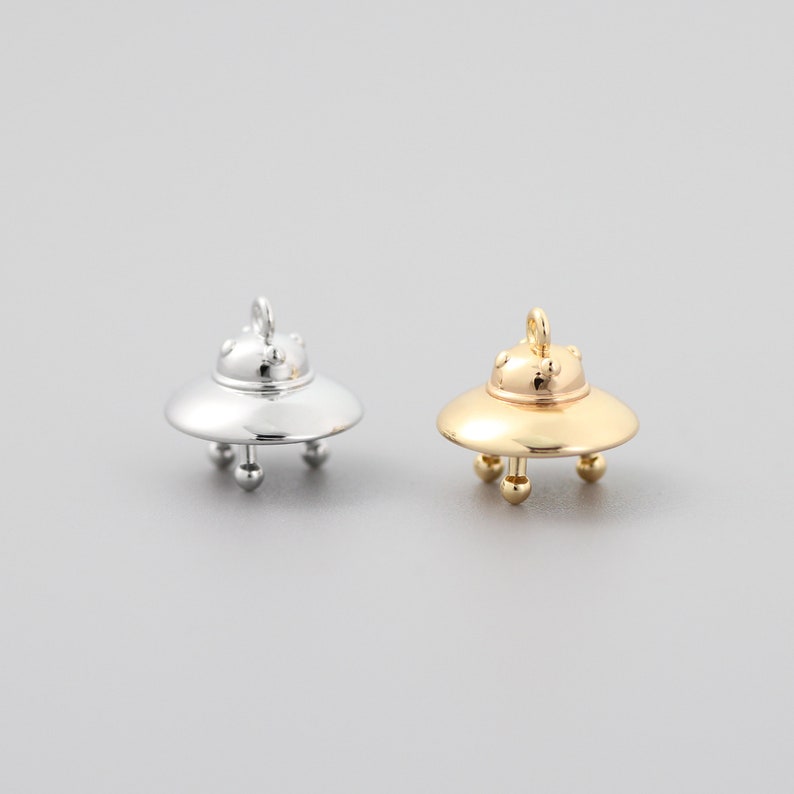 Op de afbeelding: Twee UFO-bedels in zilver en goud. De bedels zijn gevormd als vliegende schotels met een gewelfde bovenkant en drie poten. De bedels zijn perfect om een vleugje fantasie toe te voegen aan sieraden of knutselwerk.