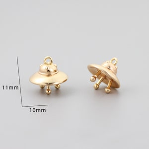 Op de afbeelding: Twee goudkleurige UFO-bedels, elk 10 mm breed en 11 mm hoog. De bedels hebben een afgeronde bovenkant en drie kleine pootjes.