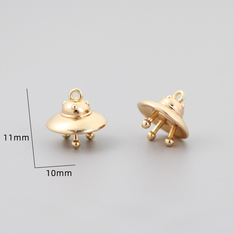 Op de afbeelding: Twee goudkleurige UFO-bedels, elk 10 mm breed en 11 mm hoog. De bedels hebben een afgeronde bovenkant en drie kleine pootjes.