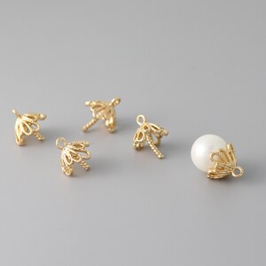 May include: Four gold-toned metal bead caps with a delicate, intricate design. The caps have a small hole at the top for threading. One cap is shown with a white pearl bead.