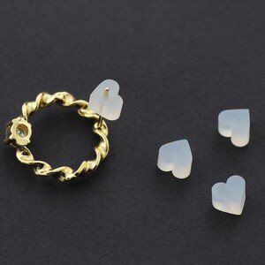 May include: Four white heart-shaped silicone earring backs. One earring back is attached to a gold-colored earring with a twisted design and a clear rhinestone.