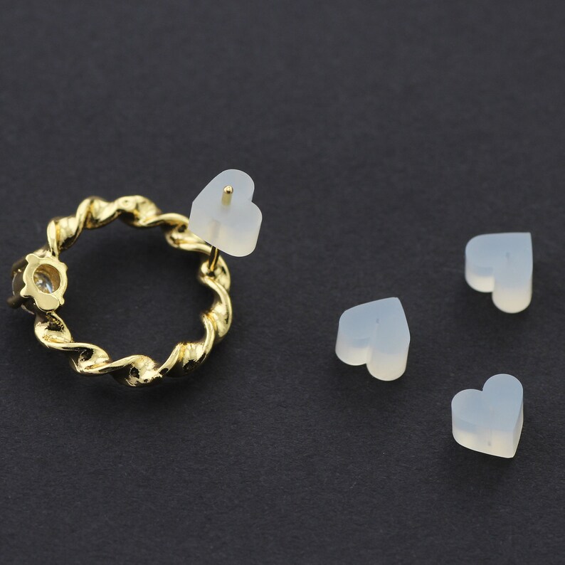 May include: Four white heart-shaped silicone earring backs. One earring back is attached to a gold-colored earring with a twisted design and a clear rhinestone.