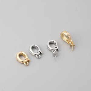Puede incluir: Cuatro cierres de joyería en tonos dorados y plateados. Se ven dos cierres dorados, uno con una fila de pequeñas piedras transparentes. También se ven dos cierres plateados. Los cierres están sobre un fondo gris.