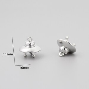 Op de afbeelding: Twee zilveren UFO-bedels, elk 10 mm breed en 11 mm hoog. De bedels hebben een afgeronde, schotelvormige vorm met een kleine lus aan de bovenkant om aan sieraden te bevestigen.