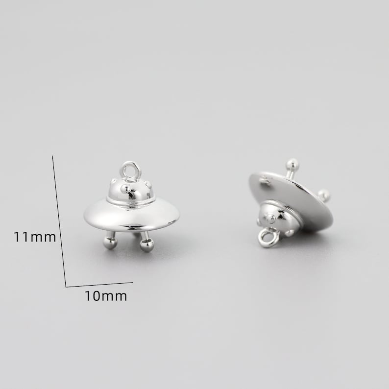 Op de afbeelding: Twee zilveren UFO-bedels, elk 10 mm breed en 11 mm hoog. De bedels hebben een afgeronde, schotelvormige vorm met een kleine lus aan de bovenkant om aan sieraden te bevestigen.
