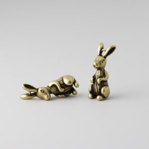 Peut inclure: Deux petites figurines de lapin en laiton antique. Un lapin est debout, l'autre est couch&eacute;. Les lapins ont des d&eacute;tails, notamment de longues oreilles et une fourrure textur&eacute;e. Le fond est de couleur claire.
