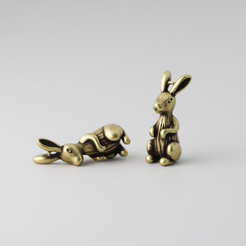 Peut inclure: Deux petites figurines de lapin en laiton antique. Un lapin est debout, l'autre est couch&eacute;. Les lapins ont des d&eacute;tails, notamment de longues oreilles et une fourrure textur&eacute;e. Le fond est de couleur claire.