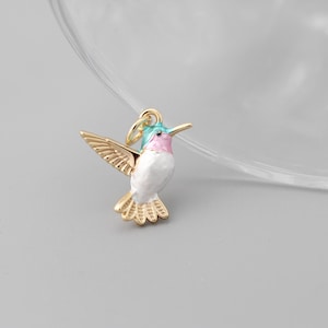 Peut inclure: Un charm en forme de colibri doré, aux ailes déployées. L'oiseau a un corps blanc, une gorge rose et une tête bleue et verte. Le charm est orné de plumes gravées et d'une petite boucle pour l'attacher.