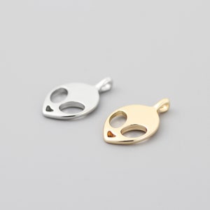 Op de afbeelding: Twee alien-hoofdcharms, een zilveren en een gouden. De charms zijn gemaakt van metaal en hebben een eenvoudig, minimalistisch ontwerp.