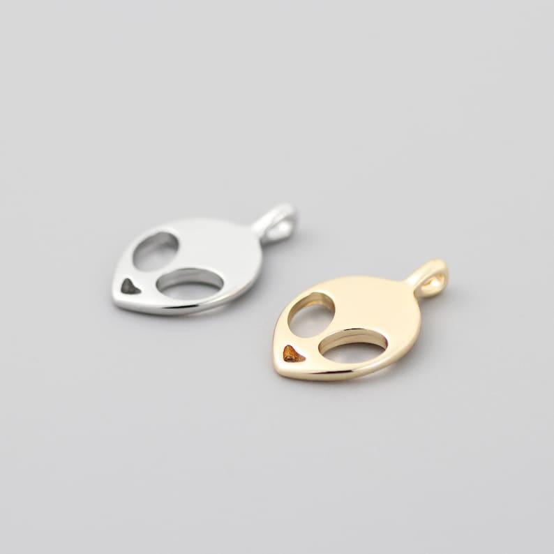 Op de afbeelding: Twee alien-hoofdcharms, een zilveren en een gouden. De charms zijn gemaakt van metaal en hebben een eenvoudig, minimalistisch ontwerp.