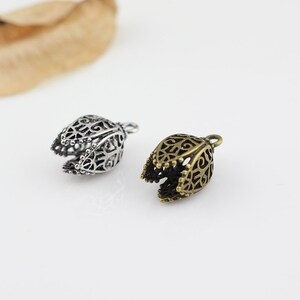 May include: Two ornate metal bell-shaped charms, one silver and one bronze, with intricate filigree designs. The charms have a loop at the top for attaching to jewelry or other crafts.
