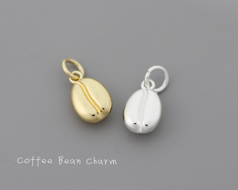 K&ouml;nnte beinhalten: Zwei Kaffebohnen-Anh&auml;nger, einer golden und einer silbern, mit einer Schlaufe zum Anbringen an einer Halskette oder einem Armband.  "Coffee Bean Charm" steht unter den Anh&auml;ngern.