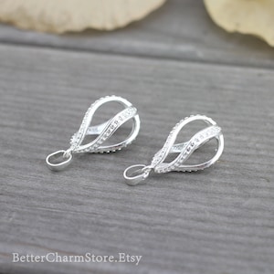 Op de afbeelding: Twee zilveren hangerbedels in druppelvorm met een delicaat, gedraaid ontwerp en een rij kleine, heldere steentjes rond de rand.