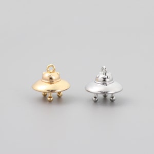 Op de afbeelding: Twee UFO-bedels in zilver en goud. De bedels zijn gevormd als vliegende schotels met een afgeronde bovenkant en drie poten. De bedels hebben een gat aan de bovenkant om aan een ketting of armband te bevestigen.