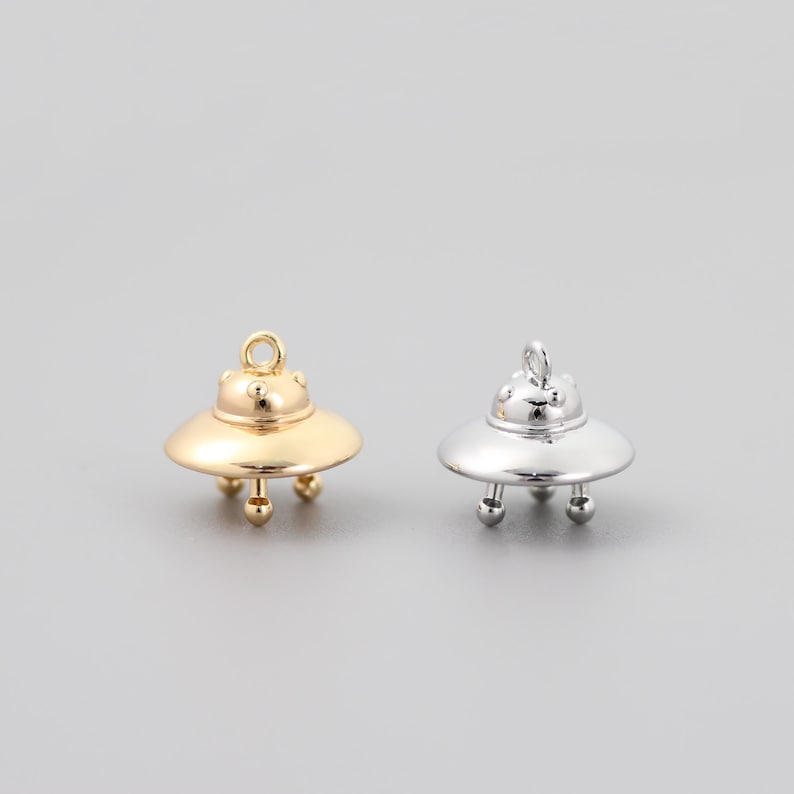 Op de afbeelding: Twee UFO-bedels in zilver en goud. De bedels zijn gevormd als vliegende schotels met een afgeronde bovenkant en drie poten. De bedels hebben een gat aan de bovenkant om aan een ketting of armband te bevestigen.