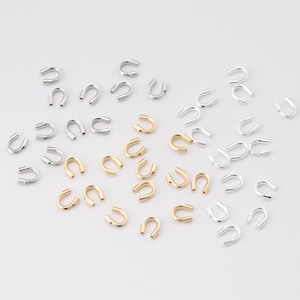 Puede incluir: Un surtido de pequeños hallazgos de joyería metálicos en forma de herradura. Los hallazgos son en tonos plateados y dorados, dispersos sobre una superficie blanca. Estos componentes se utilizan en la fabricación de joyas y manualidades.