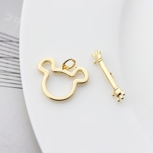 Può includere: Due ciondoli in metallo color oro. Un ciondolo ha la forma di una testa di topo dei cartoni animati con grandi orecchie. L'altro ciondolo ha la forma di una mano con un braccio lungo e sottile.