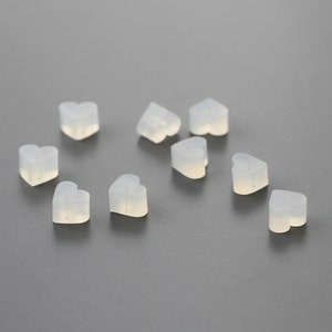 May include: Ten clear heart-shaped silicone earplugs. The earplugs are small and translucent, and they are arranged in a scattered pattern on a gray surface.