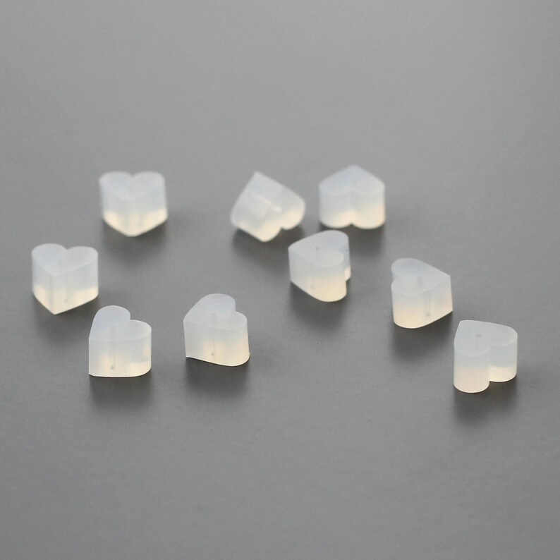 May include: Ten clear heart-shaped silicone earplugs. The earplugs are small and translucent, and they are arranged in a scattered pattern on a gray surface.