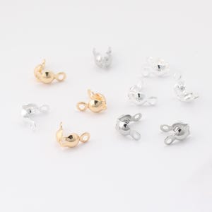 May include: A selection of small, metallic jewellery findings in gold and silver tones. These findings have a rounded shape with a loop for attaching to other jewellery components. The findings are scattered on a white surface.