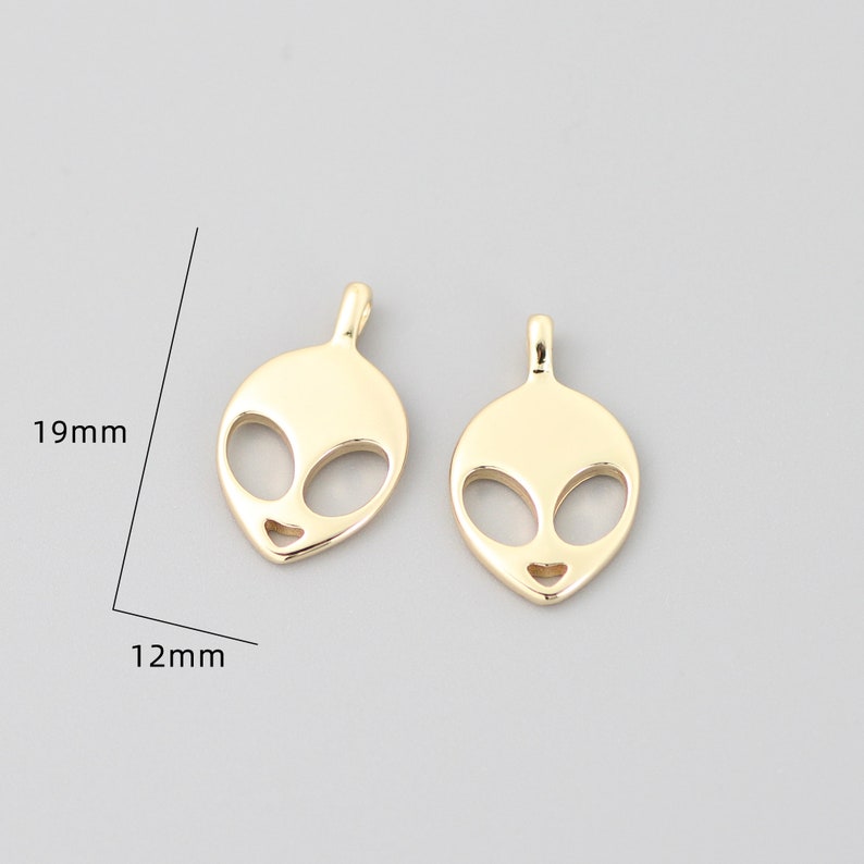 Op de afbeelding: Twee goudkleurige alien-hoofdcharms, elk 19 mm hoog en 12 mm breed. De charms zijn uitgesneden in de vorm van een alien-hoofd met grote ogen en een kleine mond.