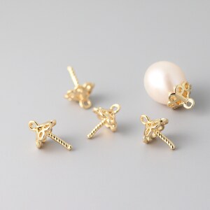 May include: Four gold-tone pearl cap settings with a delicate, intricate design. The settings have a small loop at the top and a post with a twisted design at the bottom. One pearl cap setting is shown with a white pearl.