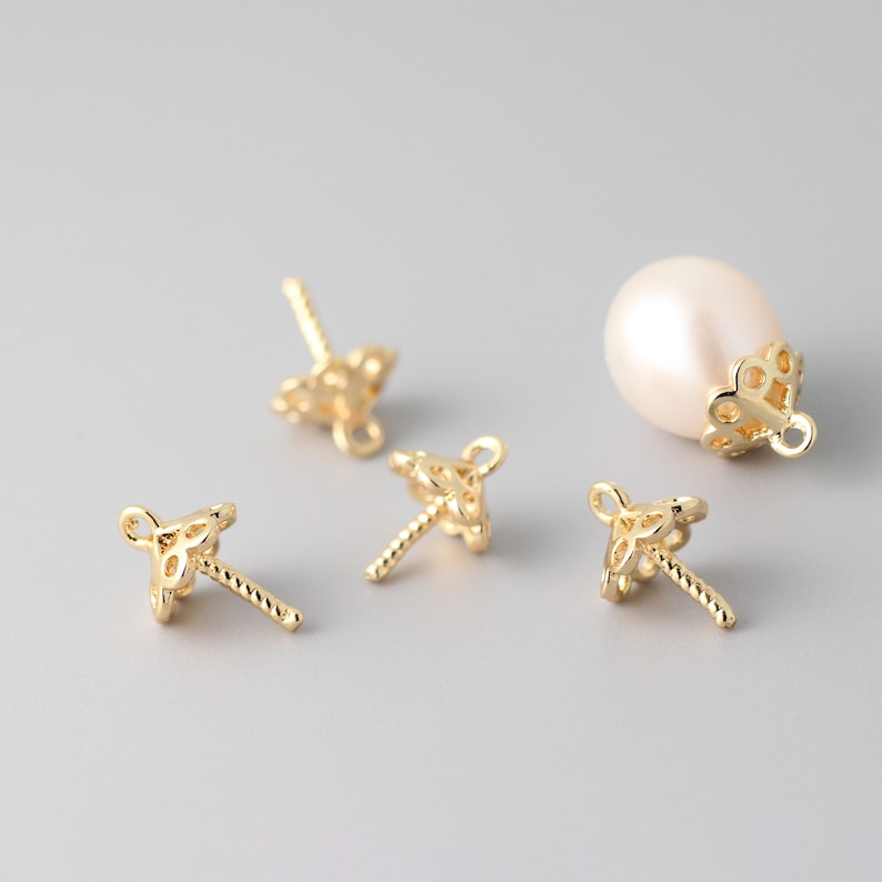 May include: Four gold-tone pearl cap settings with a delicate, intricate design. The settings have a small loop at the top and a post with a twisted design at the bottom. One pearl cap setting is shown with a white pearl.