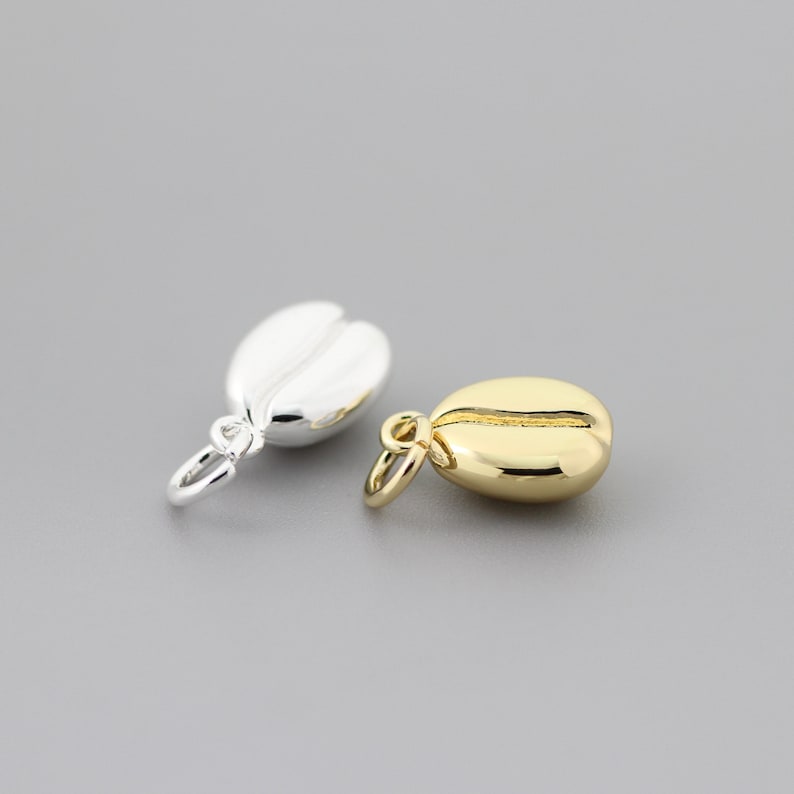 K&ouml;nnte beinhalten: Zwei Kaffebohnen-Anh&auml;nger, einer in Silber und einer in Gold, mit einer Schlaufe zum Anbringen an einer Halskette oder einem Armband.