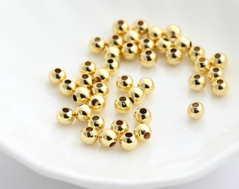 May include: A collection of small, shiny, gold-colored beads with a smooth, reflective surface. The beads are round with a hole through the center, scattered on a white surface. These beads are suitable for jewelry making and crafting.