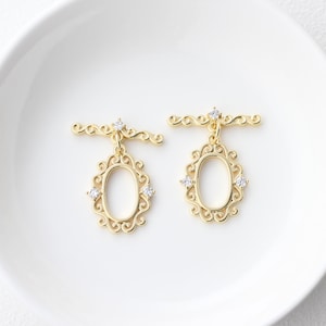 Può includere: Un paio di orecchini dorati con un delicato design a cornice ovale. Ogni orecchino presenta un piccolo cristallo trasparente al centro della cornice e due cristalli più piccoli ai lati. Gli orecchini sono attaccati a una barra dorata con un design a spirale decorativo.