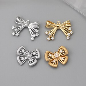 Op de afbeelding: Vier strikvormige bedels in zilver- en goudtinten. Elke bedel heeft een strikdesign met uitstralende lijnen en kleine, heldere edelstenen. De bedels worden gepresenteerd op een grijze ondergrond.