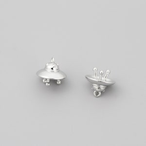 Op de afbeelding: Twee zilveren UFO-bedels. Elke bedel heeft een gedetailleerd UFO-ontwerp met een afgeronde bovenkant en een platte onderkant. E&eacute;n bedel heeft een lus aan de bovenkant om aan een ketting of armband te bevestigen. De andere bedel heeft een lus aan de onderkant.
